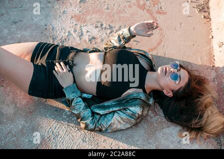 portrait of a young brunette girl with a beautiful body in black clothes and a silver jacket lying on the roof and enjoying the Stock Photo