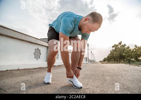 portrait of Caucasian guy in a blue t-shirt and black shorts,who ties his laces on white sports shoes before Jogging Stock Photo