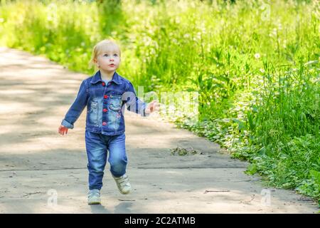 Selective focus on a blonde little girl running on an asphalt path in a park past thickets of dense green grass. Happy child plays in nature on a sunn Stock Photo