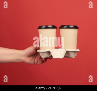 brown paper disposable cups with a plastic lid stand in the tray, female hand holds disposable tableware on a red background Stock Photo