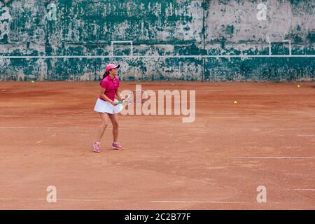 Tennis player. Side view of sportswoman practicing tennis on the court. Dressed in pink t-shirt and white skirt, in cap. Full length. Stock Photo