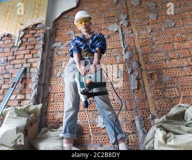 young woman in work clothes and a protective helmet stands next to a brick wall and holds a heavy ha Stock Photo