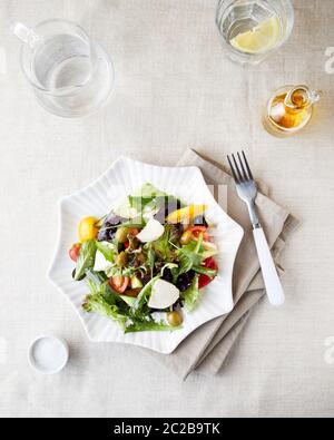 Vegetarian salad with fresh greenery, cucumbers, tomato and olives on the plate near water with lemon and olive oil bottle top view Stock Photo