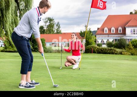 Smiling girlfriend holding flag looking at male golfer putting the golf ball in green Stock Photo