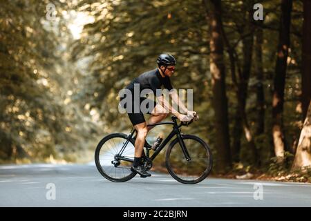 Side view of active man in sport clothing riding black bike with beautiful green trees around. Bearded cyclist in protective helmet and eyewear traini Stock Photo
