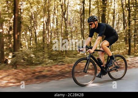 Side view of mature man with sporty body in protective helmet practising in cycling outdoors. Bearded athlete wearing mirrored glasses biking on aspha Stock Photo