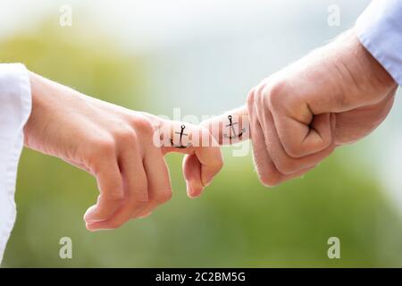 Loving Couple Holding Each Other's Finger With Black Anchor Sign Tattoo Against Blurred Background Stock Photo