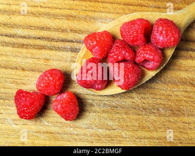 A wooden spoon full of fresh raspberries on a wooden chopping board with three raspberries spilling over Stock Photo