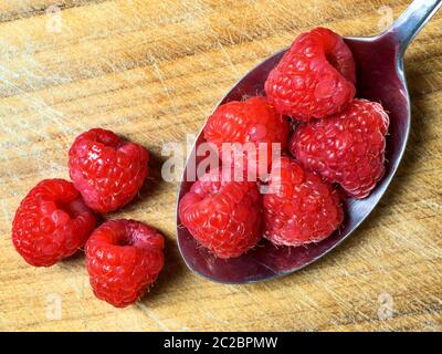 Five fresh raspberries in a stainless steel table spoon with three raspberries spilling over on a wooden chopping board Stock Photo