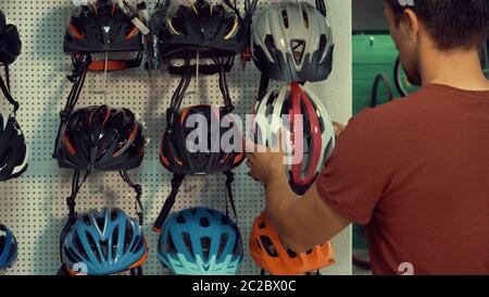 A young Caucasian man stands near a stand in a bicycle store in the head protection department. Choosing a bicycle helmet in a s Stock Photo