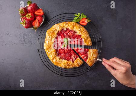 Woman's hand holds a spatula with a cut piece of cake with strawberries, mint and almonds on a dark background. Next to a pie on a stand Stock Photo