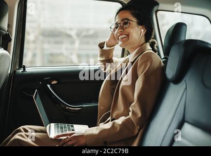 Smiling businesswoman travelling in a taxi with laptop. Woman with laptop sitting in back seat of her car looking away and smiling. Stock Photo