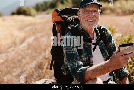 Senior male backpacker having coffee on hiking trail. Male hiker taking rest and drinking fresh coffee, looking at camera and smiling. Stock Photo