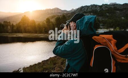 Senior man on hiking trip taking photographs of the view with a digital camera. Man hiker standing by the river in forest and taking photos. Stock Photo