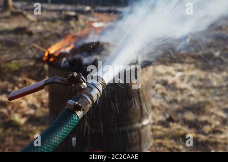 Water spraying from water hose near fire burning in old rusty barrel. Symbol of fire safety. Stock Photo