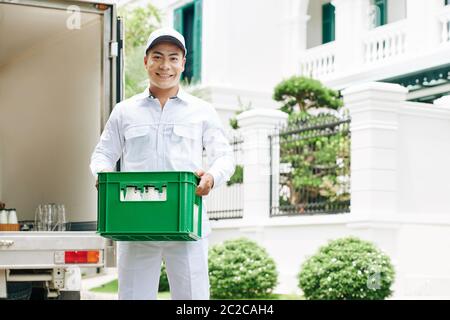 Horizontal medium long portrait shot of modern milkman wearing white clothes standing outdoors holding plastic box with milk bottles, copy space Stock Photo