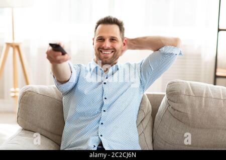 Happy guy watching TV at home, holding remote control Stock Photo