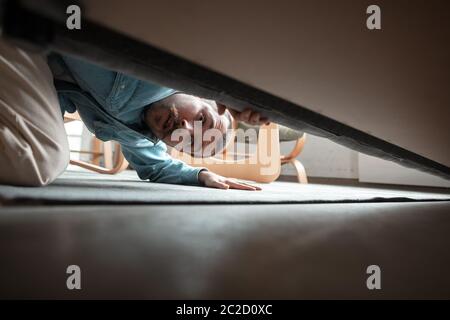 Under his bed in bedroom. Caucasian young man looking for job in unusual places at his home. Crazy, funny way to find career and going up. Concept of crisis, unemployment, finance, business. Stock Photo