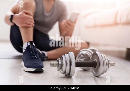 Track activity of body. Man with smart watch watches at phone, sitting on floor on mat, dumbbells near Stock Photo