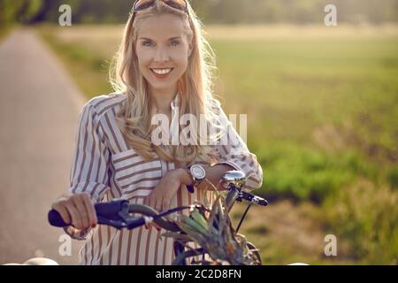 Woman using her bicycle to buy fresh produce standing on a rural road backlit by a warm glow of the sun smiling happily at the camera as she holds her Stock Photo