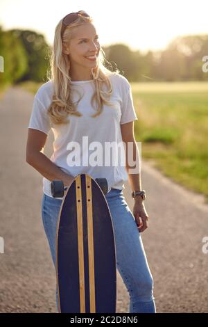 Portrait of a fit beautiful middle-aged woman with an active lifestyle smiling and looking at camera while holding a longboard on a sunny road in the Stock Photo