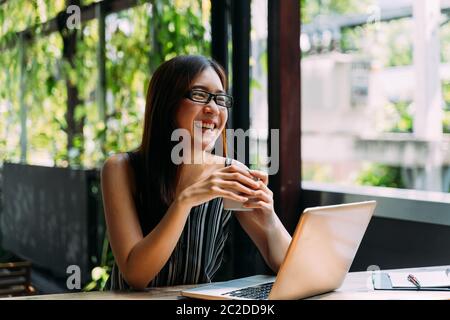 Positive Asian female student in glasses smiling and browsing laptop while sitting at table on terrace and studying on summer day Stock Photo