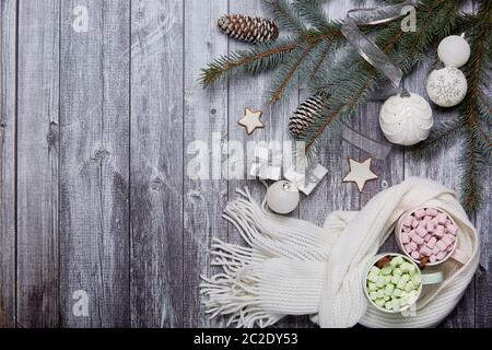 Two mugs of cocoa with marshmallow, woolen white scarf, gift boxes, fir tree branches and Christmas ornaments on grey wooden background. Top view with Stock Photo
