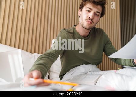 Photo of caucasian focused man with mustache doing homework while lying on bed in apartment Stock Photo