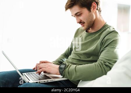 Photo of handsome young man with mustache typing on laptop while sitting on couch in apartment Stock Photo