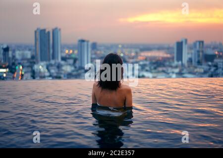 Young woman relax in swimming pool on roof top during amazing sunset and enjoy cityscape Stock Photo