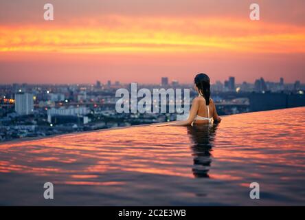 Young woman relax in swimming pool on roof top during amazing sunset and enjoy cityscape