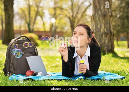 Blowing soap bubbles in the wind. Close up shot of a beautiful young student girl lying on a meadow blowing soap bubbles, taking a break from studying Stock Photo