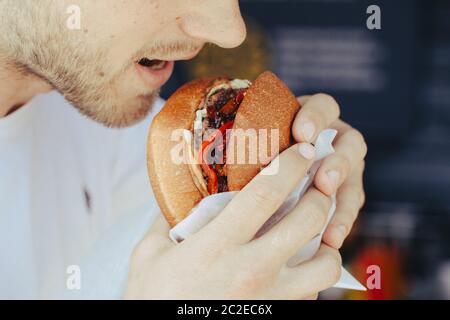 Close-up of man holds and bites hamburger, eating fast food, unhealthy food concept, toned