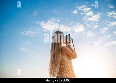 portrait of a blonde girl in a floral print dress with a vintage video camera in a grape field records video of a plane taking o Stock Photo