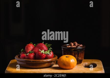 Still life with strawberries, hazelnuts and a tangerine with ceramic containers on a kitchen wooden board Stock Photo