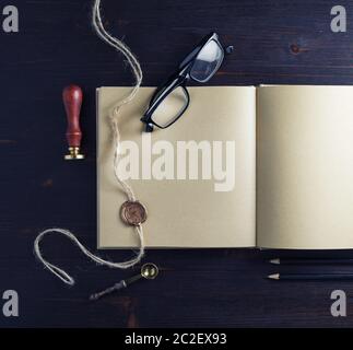 Opened kraft paper brochure and stationery on wood table background. Book, glasses, stamp, sealing wax and pencils. Flat lay. Stock Photo