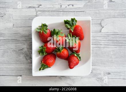 Fresh juicy red strawberries with green leaves lying on a white plate. Light grey wooden table background. Stock Photo