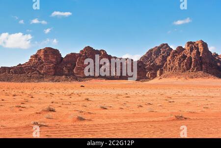 Red dusty desert with large rock massif and blue sky in background, small off road vehicle in foreground for scale. Typical landscape of Wadi Rum Stock Photo