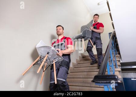 Portrait Of A Young Male Movers In Uniform Carrying Chairs Walking Downward On Staircase Stock Photo
