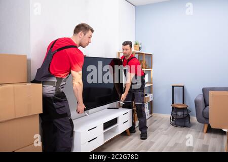 Two Male Professional Movers In Uniform Unloading LCD Television In The Living Room Stock Photo