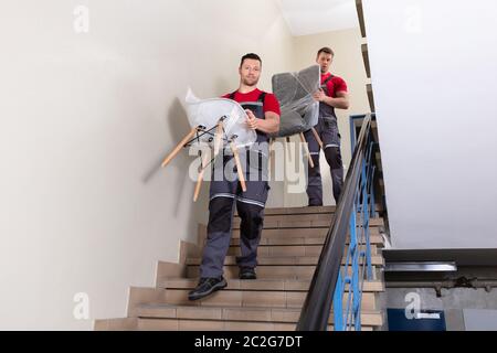 Portrait Of A Young Male Movers In Uniform Carrying Chairs Walking Downward On Staircase Stock Photo