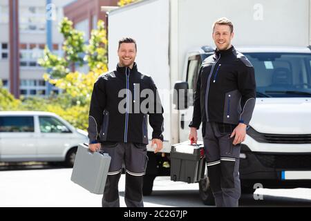 Portrait Of Smiling Two Male Electrician Carrying Tool Boxes Standing Against Truck Looking At Camera Stock Photo