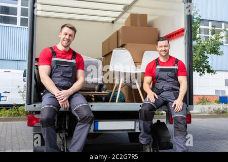 Portrait Of Smiling Movers Sitting In Van With Stack Of Cardboard Boxes And Furniture Stock Photo