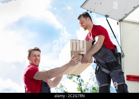 Male Movers Unloading And Passing A Cardboard Box To His Assistant In Moving Van Stock Photo