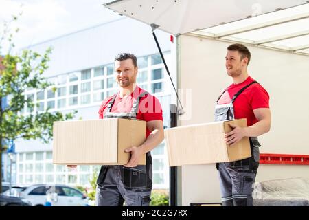 Happy Young Male Movers Holding Cardboard Boxes Standing In Moving Van Stock Photo