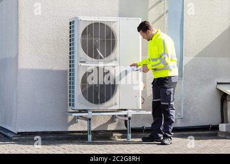 Young Male Technician Wearing Safety Jacket Looking At Air Conditioner Unit Making Notes Stock Photo
