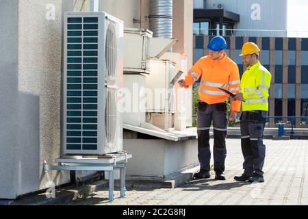 Two Electricians Men Wearing Safety Jackets Checking Air Conditioning Unit On Building Rooftop Stock Photo