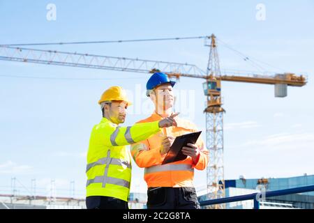Two Young Male Engineers Standing In Front Of Mechanical Crane Working On The Construction Site Stock Photo