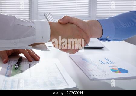 Close-up Of Two Businessmen Shaking Hands Over Financial Graph Report On White Desk In Office Stock Photo
