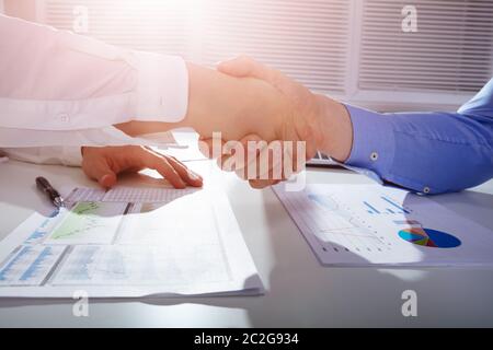 Close-up Of Two Businessmen Shaking Hands Over Financial Graph Report On White Desk In Office Stock Photo